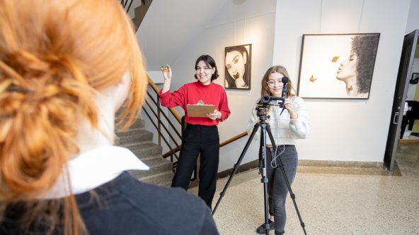 Photo de deux élèves se préparant à en filmer une autre avec une caméra et un trépied.