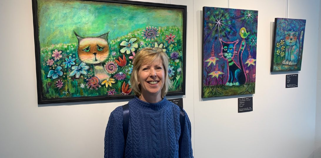 L'artiste Caroline Singler pose devant trois de ses oeuvres, dont une qui représente un chat dans un champ de fleurs.